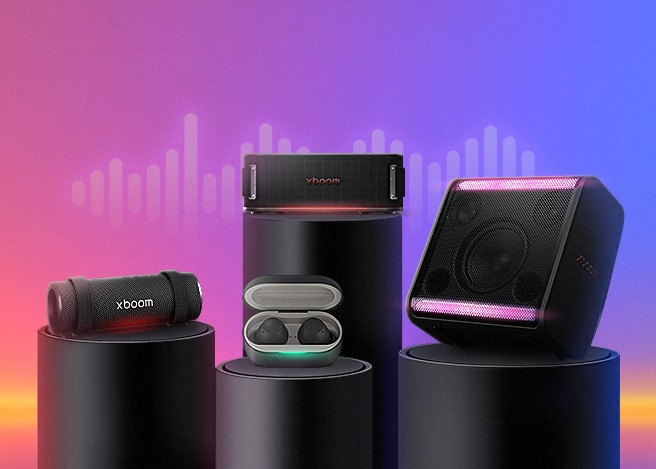 그라데이션 배경 위에 다양한 형태의 LG xboom 스피커 제품들이 원형 받침 위에 전시되어 있으며, 중앙에는 무선 이어폰과 조명이 켜진 스피커가 함께 배치되어 있습니다.